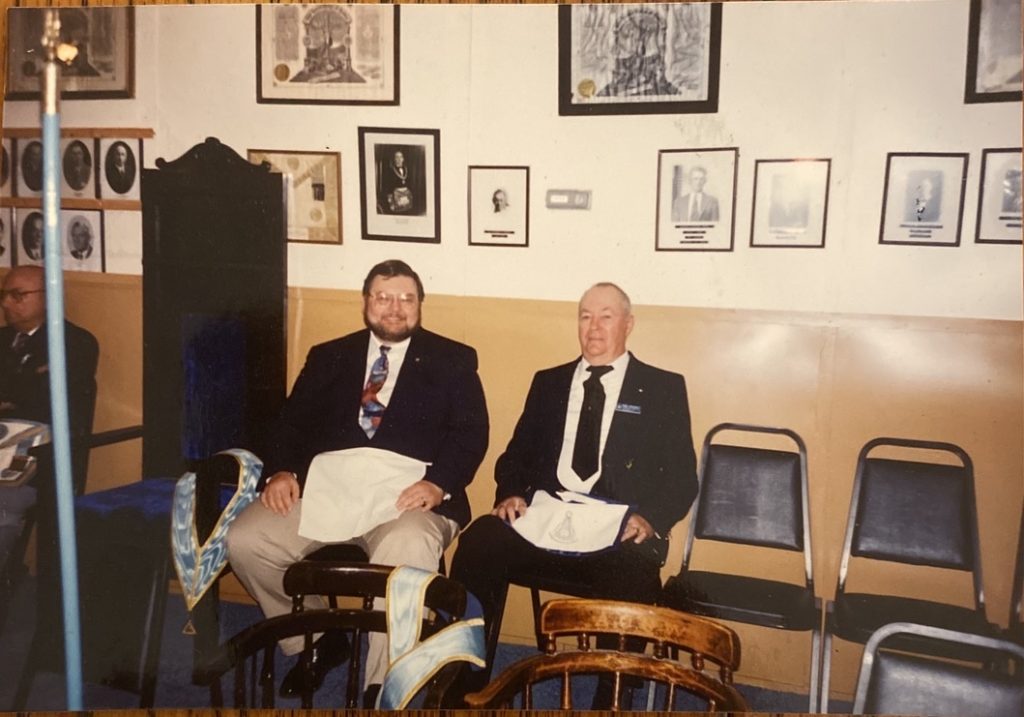 Two men wearing white Masons aprons sit in a room with photos hanging on the wall behind them.
