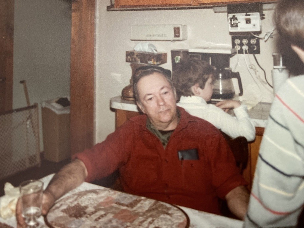 A man sits at a dining table with a quilted placemat in front of him and a glass of water in his right hand
